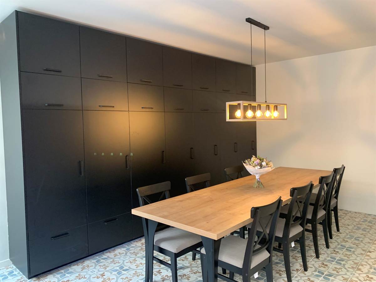 Salle à manger avec une grande table en chêne, des chaises noires et un mur de rangements sombres.